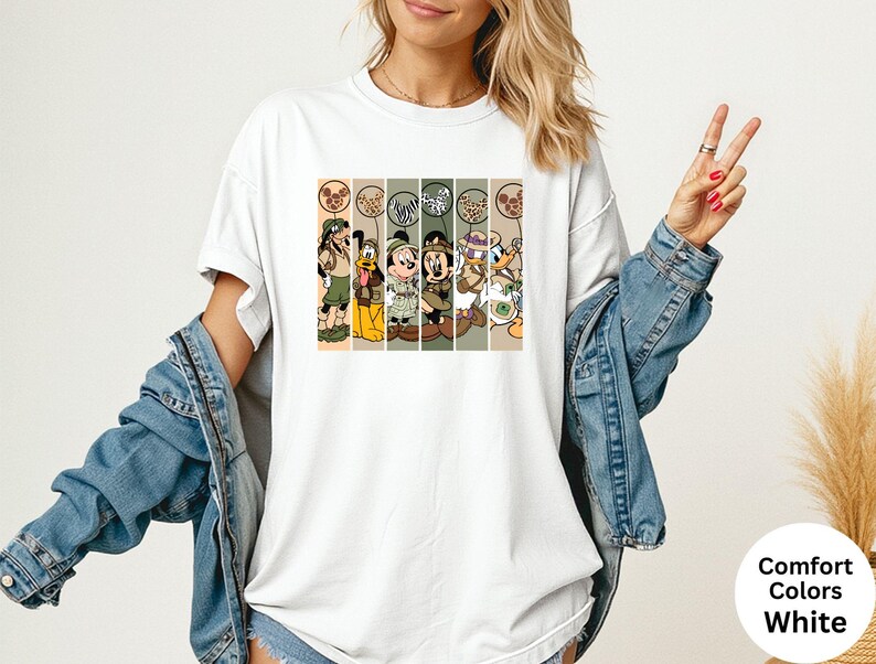 May include: White t-shirt featuring a graphic of Disney characters in safari outfits. The design includes Goofy, Donald Duck, Mickey Mouse, and Minnie Mouse. The shirt has the words "Comfort Colors White" on the bottom right.