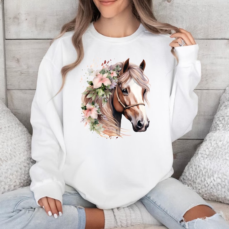 Peut inclure: Sweat-shirt blanc avec une illustration &agrave; l'aquarelle d'une t&ecirc;te de cheval orn&eacute;e de fleurs roses et de verdure. Le cheval a une crini&egrave;re brun clair et un licol marron. Le motif est centr&eacute; sur le devant du sweat-shirt.