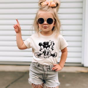 May include: A young girl wearing a white t-shirt with a black and white cartoon image of Mickey Mouse and Minnie Mouse. She is wearing denim shorts and sunglasses.
