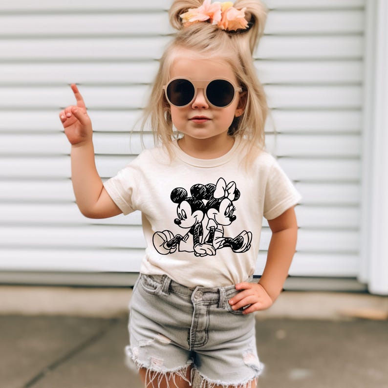 May include: A young girl wearing a white t-shirt with a black and white cartoon image of Mickey Mouse and Minnie Mouse. She is wearing denim shorts and sunglasses.