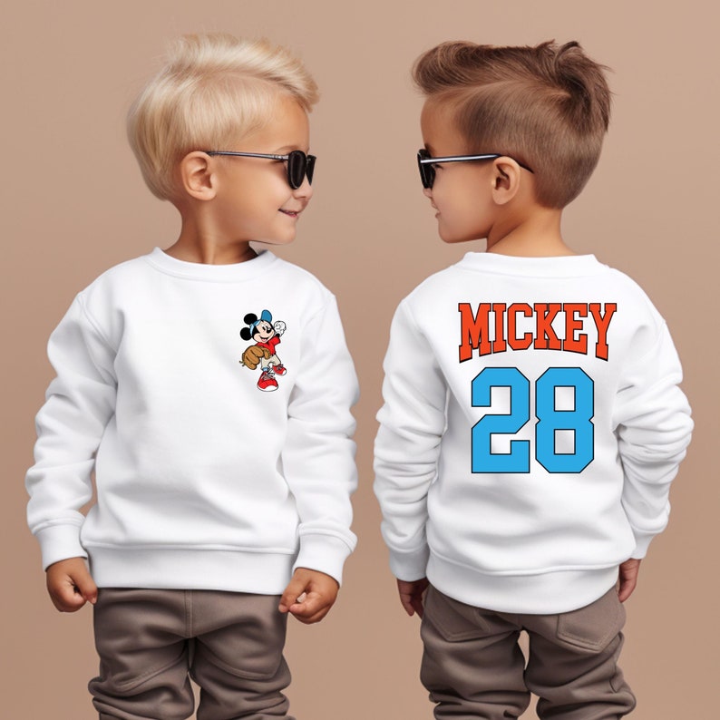 May include: Two children wearing white sweatshirts with a Mickey Mouse design. One sweatshirt has a small Mickey Mouse graphic on the front, while the other has "MICKEY 28" printed on the back in blue and orange.