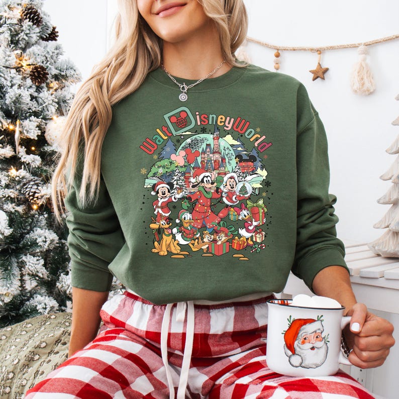 May include: Green sweatshirt with a colorful graphic featuring Disney characters in holiday attire, including Mickey Mouse, Goofy, and Donald Duck, with the words "Walt Disney World". A white mug with a Santa Claus design is also visible.