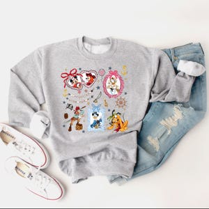 May include: A gray sweatshirt with a colorful print of Disney characters in frames. The characters include Mickey Mouse, Minnie Mouse, Donald Duck, Goofy, and Pluto. The print also includes hearts, stars, and anchors.