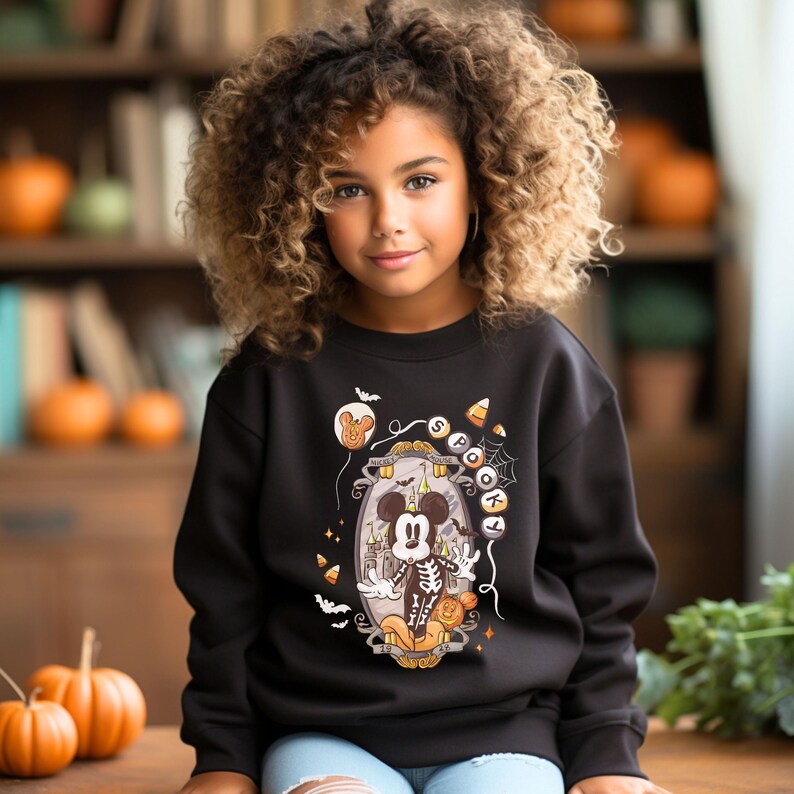 May include: A black sweatshirt featuring a Halloween-themed graphic of Mickey Mouse as a skeleton inside a decorative frame. The design includes bats, candy corn, and the word "Spooky". The sweatshirt is worn by a young person with curly hair.