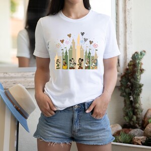 May include: White t-shirt with a graphic of Mickey Mouse and friends in front of a castle silhouette with fireworks and balloons. The castle is in shades of green and brown.