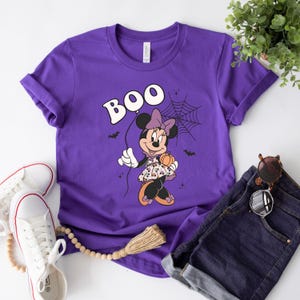 May include: Purple t-shirt with a graphic of Minnie Mouse dressed in a Halloween costume holding a pumpkin and a spider web. The text "BOO" is above Minnie Mouse.