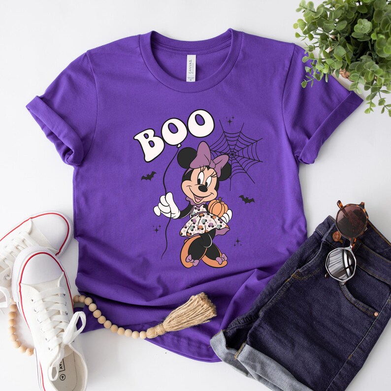 May include: Purple t-shirt with a graphic of Minnie Mouse dressed in a Halloween costume holding a pumpkin and a spider web. The text "BOO" is above Minnie Mouse.