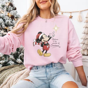 May include: A light pink sweatshirt featuring a cartoon Mickey Mouse in a Santa hat and scarf, pointing at a star. The text "Believe in the wonder of Christmas" is printed on the front. The sweatshirt is a festive holiday item.
