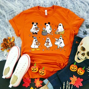 May include: Orange t-shirt featuring a graphic of various Disney characters dressed as ghosts for Halloween. The characters include Mickey Mouse, Minnie Mouse, Donald Duck, Goofy, Pluto, and a ghost holding a cauldron.