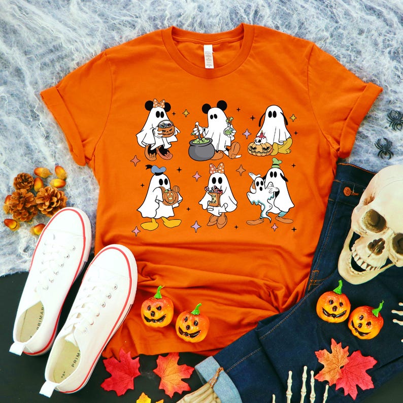 May include: Orange t-shirt featuring a graphic of various Disney characters dressed as ghosts for Halloween. The characters include Mickey Mouse, Minnie Mouse, Donald Duck, Goofy, Pluto, and a ghost holding a cauldron.