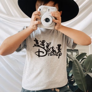 May include: A gray t-shirt with a black silhouette of Mickey Mouse and friends walking in a line with the word "DISNEY" underneath. A child is wearing the shirt and holding a white Instax camera.