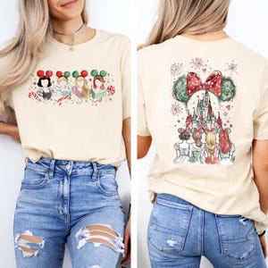 May include: A beige t-shirt featuring a watercolor design of Disney princesses wearing holiday-themed Mickey Mouse ears. The back of the shirt shows a castle and more princesses with a large bow and fireworks.