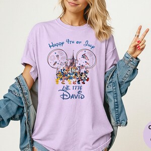 May include: A light purple t-shirt with a graphic featuring Mickey Mouse ears, a castle, fireworks, and Disney characters. The text reads "Happy 4th of July" and "Est. 1776 David". The shirt is a Comfort Colors Orchid.