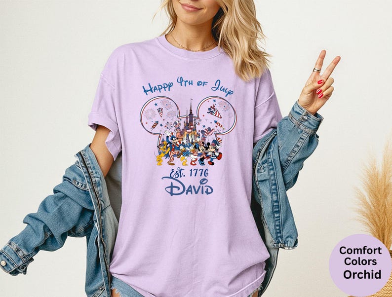 May include: A light purple t-shirt with a graphic featuring Mickey Mouse ears, a castle, fireworks, and Disney characters. The text reads "Happy 4th of July" and "Est. 1776 David". The shirt is a Comfort Colors Orchid.