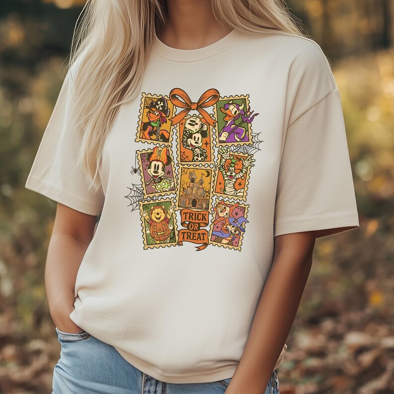 May include: Beige t-shirt featuring a colorful Halloween-themed graphic. The graphic includes postage stamp-style images of cartoon characters, a castle, and the words "Trick or Treat" on a ribbon. A large orange bow is at the top of the graphic.