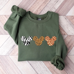 May include: An olive green sweatshirt featuring three Mickey Mouse head shapes with animal print designs: zebra, leopard, and giraffe. The sweatshirt has long sleeves and a crew neck. The fabric appears soft and comfortable.
