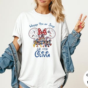 Pode incluir: Camiseta branca com um design patri&oacute;tico com orelhas do Mickey, um la&ccedil;o vermelho e o texto "Happy 4th of July". O design inclui fogos de artif&iacute;cio e o texto "Est. 1776 Olivia" em azul. A camisa &eacute; um estilo de cores confort&aacute;veis.