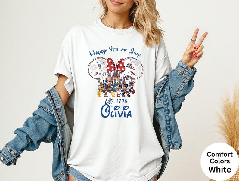 Pode incluir: Camiseta branca com um design patri&oacute;tico com orelhas do Mickey, um la&ccedil;o vermelho e o texto "Happy 4th of July". O design inclui fogos de artif&iacute;cio e o texto "Est. 1776 Olivia" em azul. A camisa &eacute; um estilo de cores confort&aacute;veis.