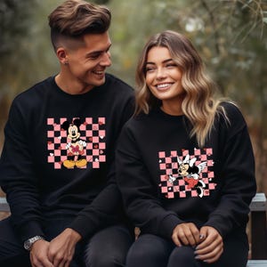 Pode incluir: Um casal vestindo su&eacute;teres pretos com um padr&atilde;o xadrez rosa e branco e uma imagem de desenho animado do Mickey Mouse e da Minnie Mouse. O Mickey Mouse est&aacute; segurando um cora&ccedil;&atilde;o e a Minnie Mouse est&aacute; segurando uma flecha.