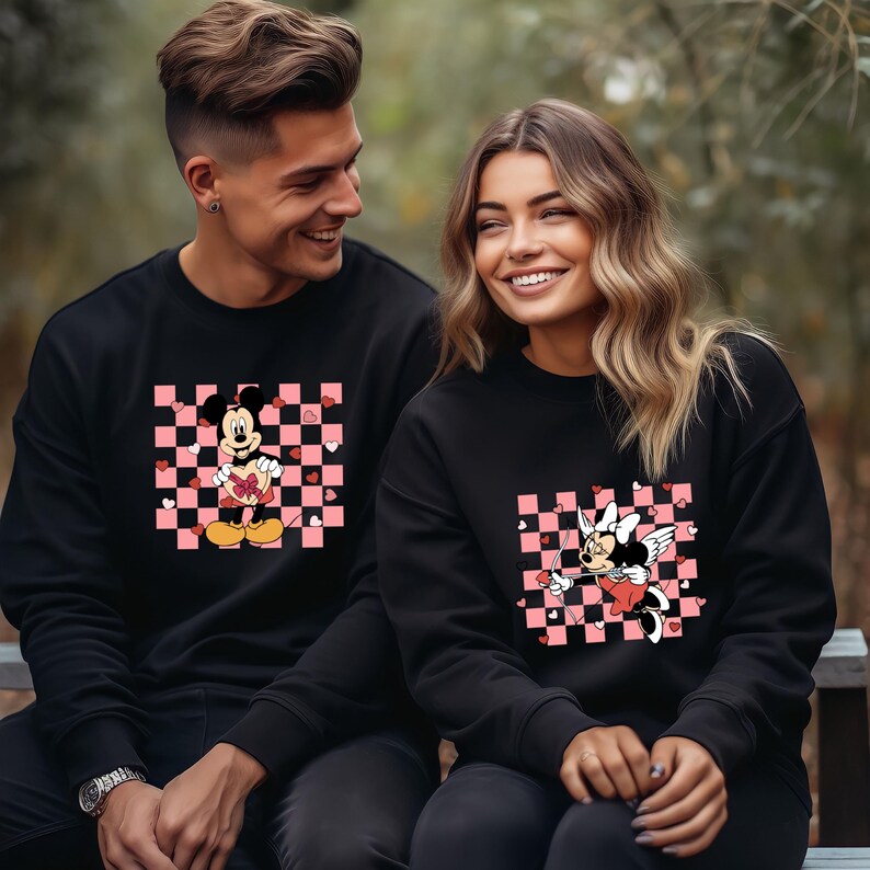 Pode incluir: Um casal vestindo su&eacute;teres pretos com um padr&atilde;o xadrez rosa e branco e uma imagem de desenho animado do Mickey Mouse e da Minnie Mouse. O Mickey Mouse est&aacute; segurando um cora&ccedil;&atilde;o e a Minnie Mouse est&aacute; segurando uma flecha.