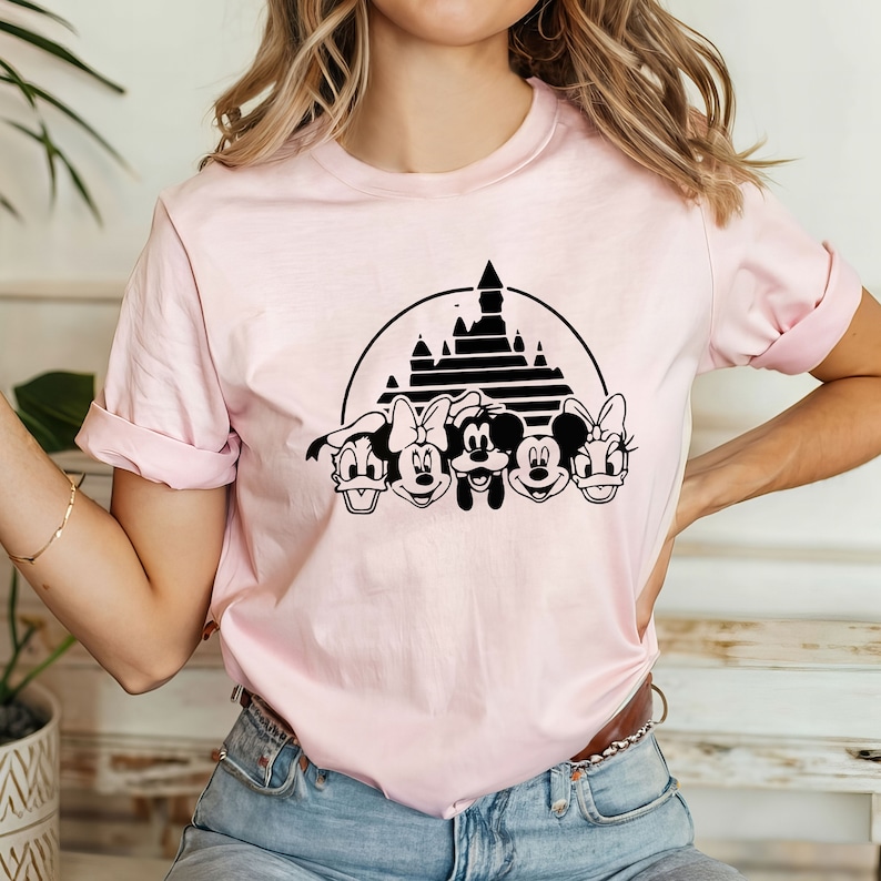 May include: A pink t-shirt with a black graphic of Mickey Mouse, Minnie Mouse, Donald Duck, Goofy, and Pluto in front of the Cinderella Castle.