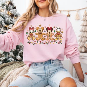 May include: A light pink sweatshirt featuring gingerbread versions of Disney characters: Daisy Duck, Mickey Mouse, Minnie Mouse, and Donald Duck. The design includes a string of colorful Christmas lights at the bottom. The sweatshirt is a festive holiday item.