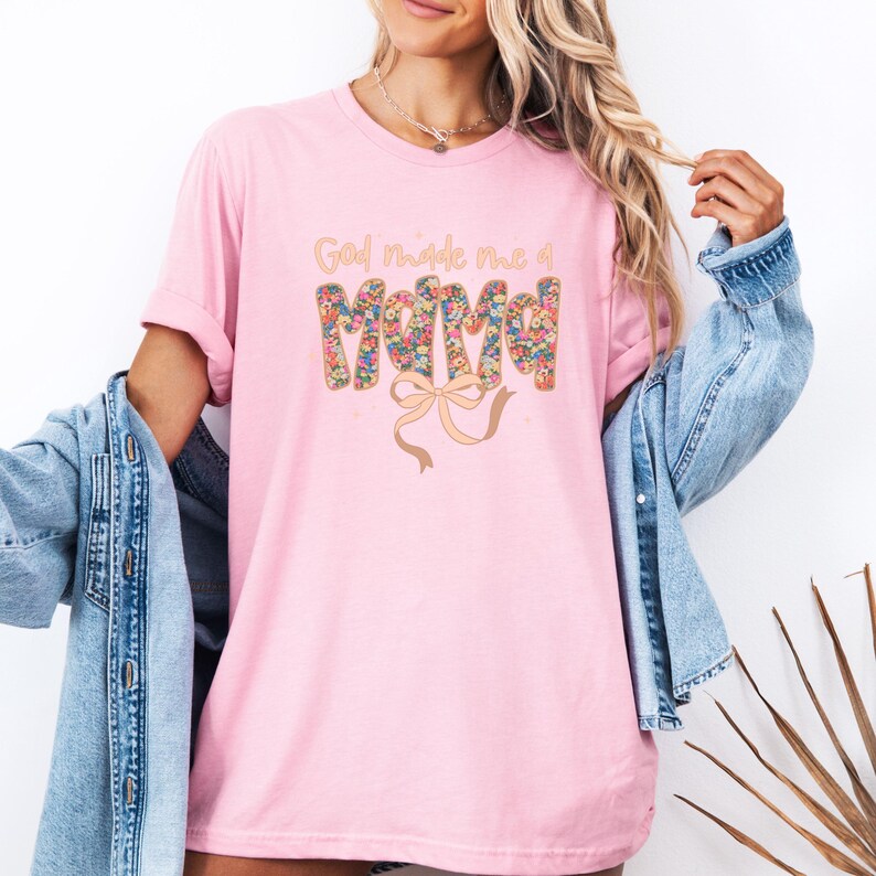 Pu&ograve; includere: T-shirt rosa chiaro con la scritta dorata "God made me a Mama". La parola "Mama" &egrave; decorata con un motivo floreale. Un disegno a nastro beige &egrave; sotto la parola "Mama".
