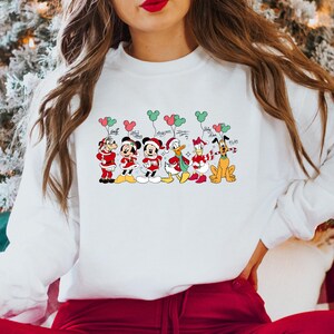 May include: White crewneck sweatshirt featuring a festive graphic of Disney characters in holiday attire, holding Mickey Mouse-shaped balloons. The characters include Mickey, Minnie, Donald, Daisy, Goofy, and Pluto, with their signatures.