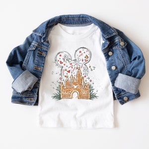 May include: White t-shirt with a gingerbread castle design. The castle is decorated with red and green candy canes, red ornaments, and a Mickey Mouse head with fireworks. The design is surrounded by green holly leaves and red berries.