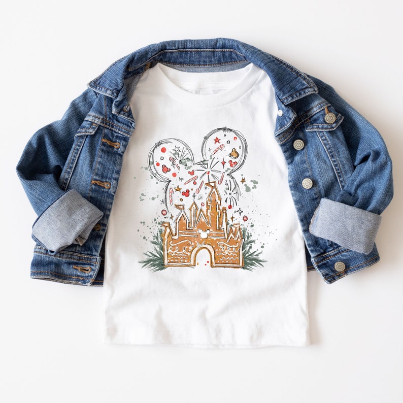 May include: White t-shirt with a gingerbread castle design. The castle is decorated with red and green candy canes, red ornaments, and a Mickey Mouse head with fireworks. The design is surrounded by green holly leaves and red berries.