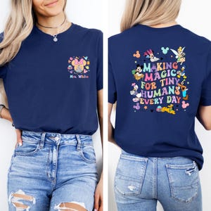 May include: Navy blue t-shirt with a colorful graphic on the front and back. The front features a Minnie Mouse design with the text "Mrs. White." The back displays the phrase "MAKING MAGIC FOR TINY HUMANS EVERY DAY" with Disney characters.