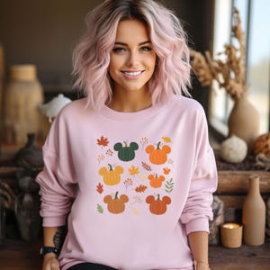May include: A light pink sweatshirt featuring a fall-themed design. The design includes pumpkins in various colors, some with Mickey Mouse ears, along with autumn leaves and floral accents. The sweatshirt has long sleeves.