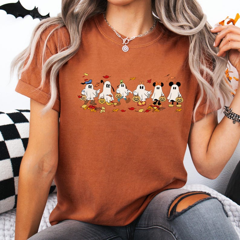 May include: A burnt orange t-shirt featuring a Halloween-themed graphic. The graphic depicts six cartoon ghosts, each with unique hats and accessories, holding pumpkin-shaped treat buckets. Autumn leaves and pumpkins are scattered around the ghosts.