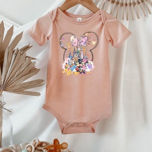 May include: A peach-colored baby onesie with a graphic of Mickey Mouse and friends in front of Cinderella's castle. The graphic is surrounded by a Mickey Mouse head shape with pink and purple butterflies.