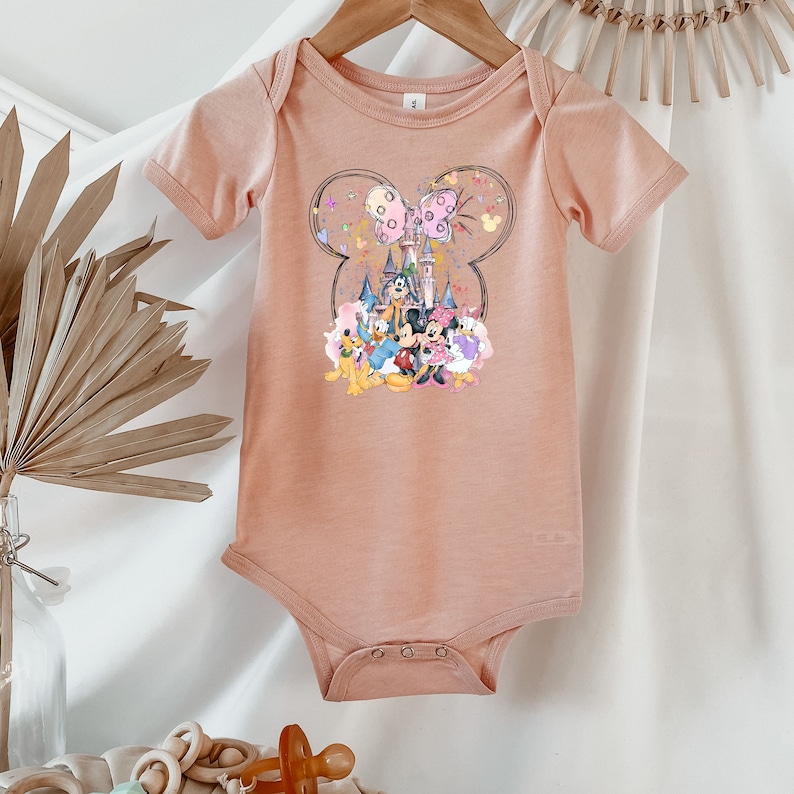 May include: A peach-colored baby onesie with a graphic of Mickey Mouse and friends in front of Cinderella's castle. The graphic is surrounded by a Mickey Mouse head shape with pink and purple butterflies.