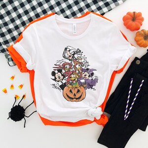 May include: White t-shirt with a colorful Halloween-themed graphic featuring cartoon characters, including Mickey Mouse, Donald Duck, and Minnie Mouse, surrounding a carved pumpkin. The shirt is folded with orange trim, and black pants are visible.