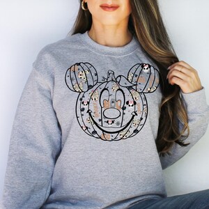 May include: A grey sweatshirt featuring a black and white outline of a Mickey Mouse pumpkin. The pumpkin is filled with smaller images of Mickey Mouse and Minnie Mouse in various Halloween costumes.