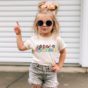 Peut inclure: Une jeune fille portant un t-shirt cr&egrave;me avec un imprim&eacute; de princesses Disney dans leurs robes. La fille porte un short en jean et des lunettes de soleil rondes.