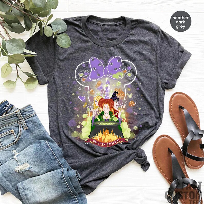 May include: A heather dark grey t-shirt with a graphic of the Sanderson sisters from the movie Hocus Pocus. The graphic features the sisters in a cauldron with a Mickey Mouse head silhouette behind them. The text "Hocus Pocus" is written below the graphic.