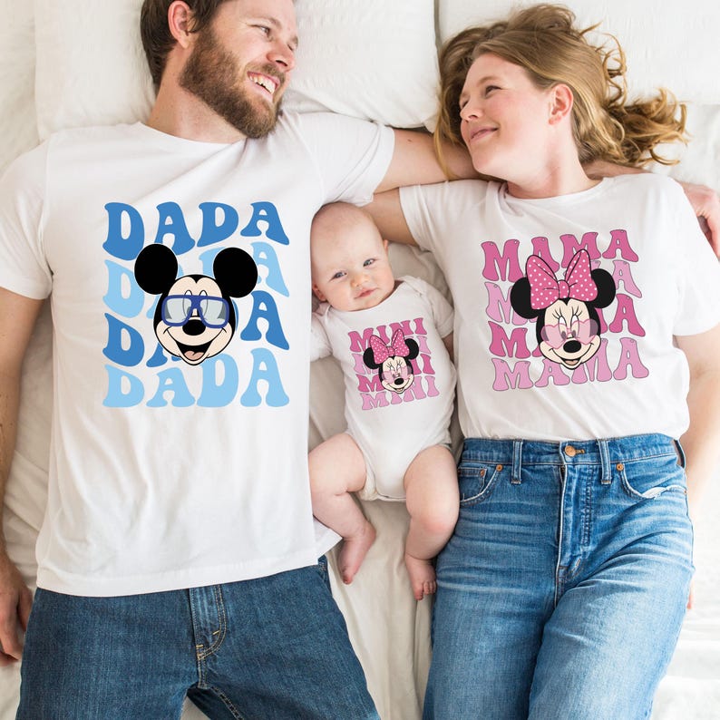 May include: A family of three wearing matching white t-shirts with a Mickey Mouse and Minnie Mouse design. The shirts say "Dada", "Mama", and "Mini" in a retro font.