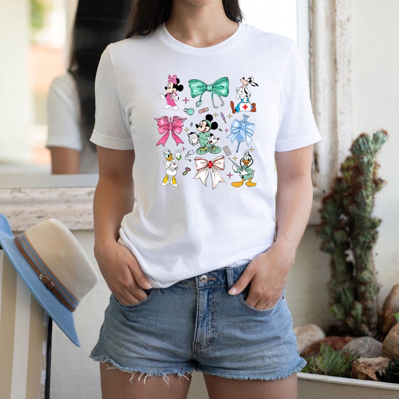 Pu&ograve; includere: T-shirt bianca con personaggi dei cartoni animati come Minnie Mouse, Topolino e Paperino, ciascuno raffigurato con simboli medici e fiocchi. Il design include fiocchi rosa, verdi e bianchi, oltre a segni di assistenza medica.