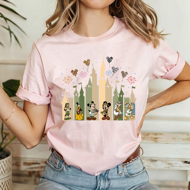 May include: Pink t-shirt with a graphic of Mickey Mouse and friends in front of a castle with balloons and fireworks. The characters are dressed in safari outfits.