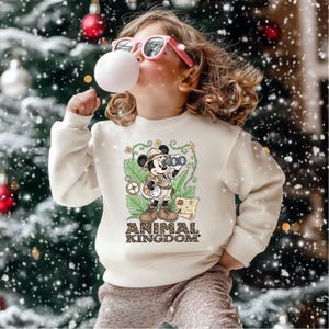 May include: Cream-colored sweatshirt featuring a graphic of Mickey Mouse in safari attire, holding binoculars, with the text "ANIMAL KINGDOM." The design includes a compass, map, and jungle foliage. The sweatshirt is worn by a person wearing pink sunglasses.