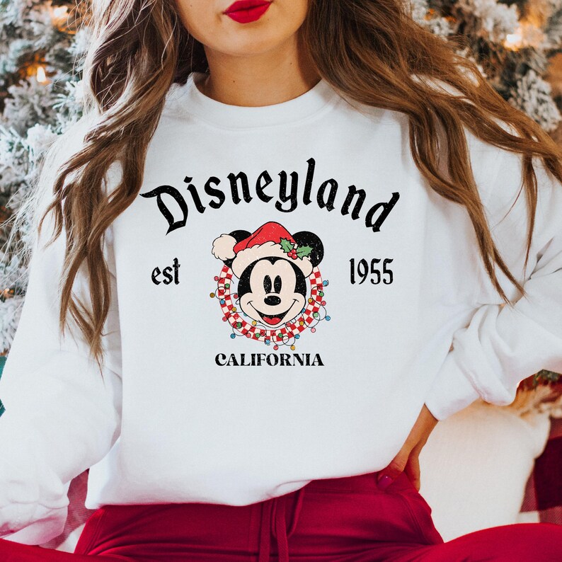 Puede incluir: Sudadera blanca con un logotipo de Disneyland de estilo vintage que presenta a Mickey Mouse con un gorro de Pap&aacute; Noel y rodeado de luces navide&ntilde;as. El texto dice "Disneyland est 1955 California".