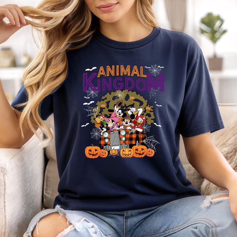 May include: Navy blue t-shirt with the text "ANIMAL KINGDOM" in purple and orange. The shirt features a graphic of Disney characters on a truck with pumpkins and a tree. Spiderwebs and bats are also included in the design.