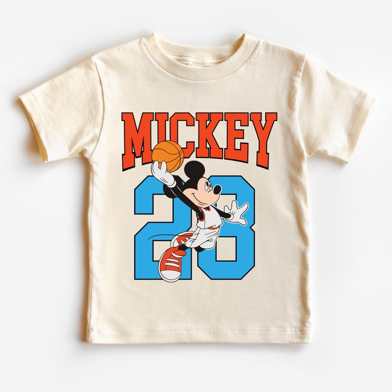以下が含まれることがあります： クリーム色のTシャツに、バスケットボールをしているミッキーマウスの漫画の画像がプリントされています。画像の上に「MICKEY」という文字が赤でプリントされ、画像の下に「23」という数字が青でプリントされています。