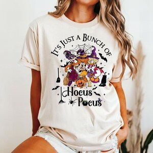 May include: A white t-shirt with a black graphic print of three cartoon characters dressed as witches. The text "It's Just A Bunch Of Hocus Pocus" is printed on the shirt.