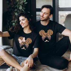 May include: A couple wearing black t-shirts with a graphic of Mickey Mouse and Minnie Mouse ears. The ears are covered in a leopard print pattern. Minnie Mouse's bow is pink with white polka dots.