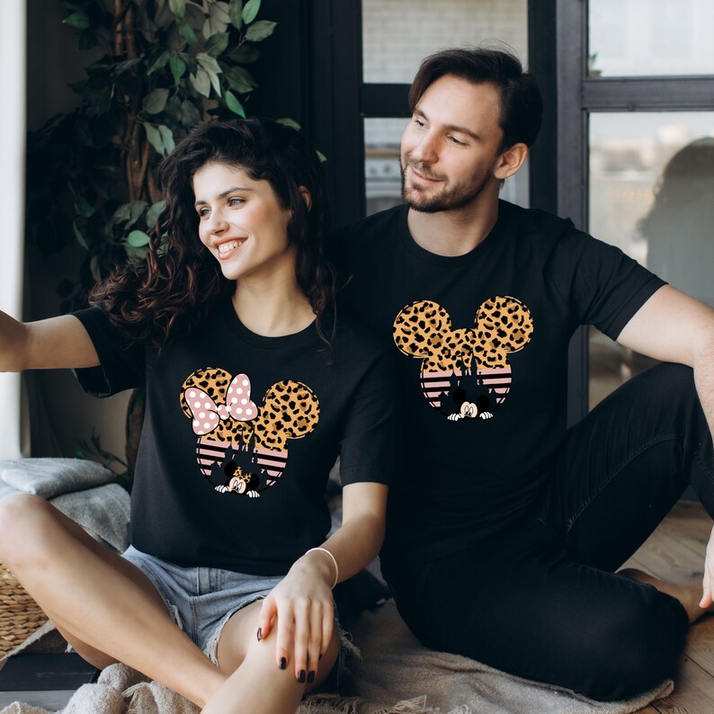 May include: A couple wearing black t-shirts with a graphic of Mickey Mouse and Minnie Mouse ears. The ears are covered in a leopard print pattern. Minnie Mouse's bow is pink with white polka dots.