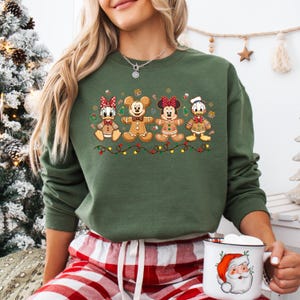May include: A green sweatshirt featuring gingerbread versions of Disney characters: Daisy Duck, Mickey Mouse, Minnie Mouse, and Donald Duck. The design includes a string of lights and festive accents, perfect for the holiday season.